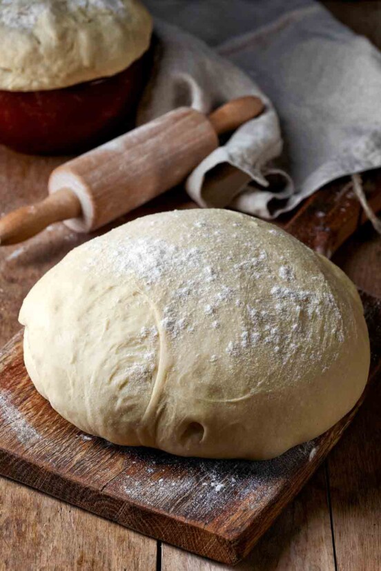 A boule of dough resting on a wooden cutting board with a rolling pin behind it.