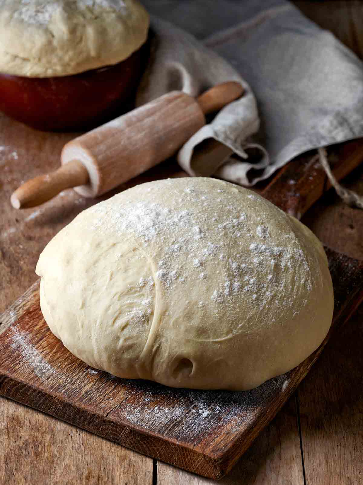 A boule of dough resting on a wooden cutting board with a rolling pin behind it.