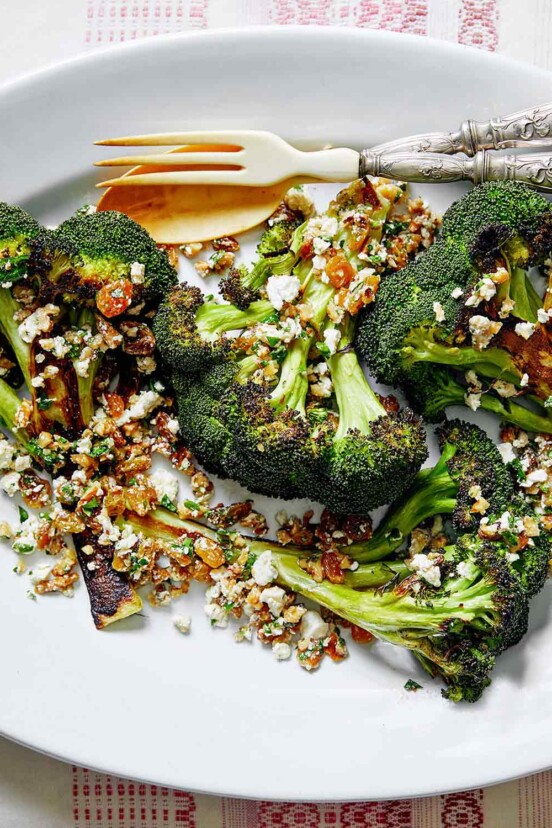 A white oval plate topped with broccoli with walnuts, raisins, and ricotta salata with a fork and spoon on the side.