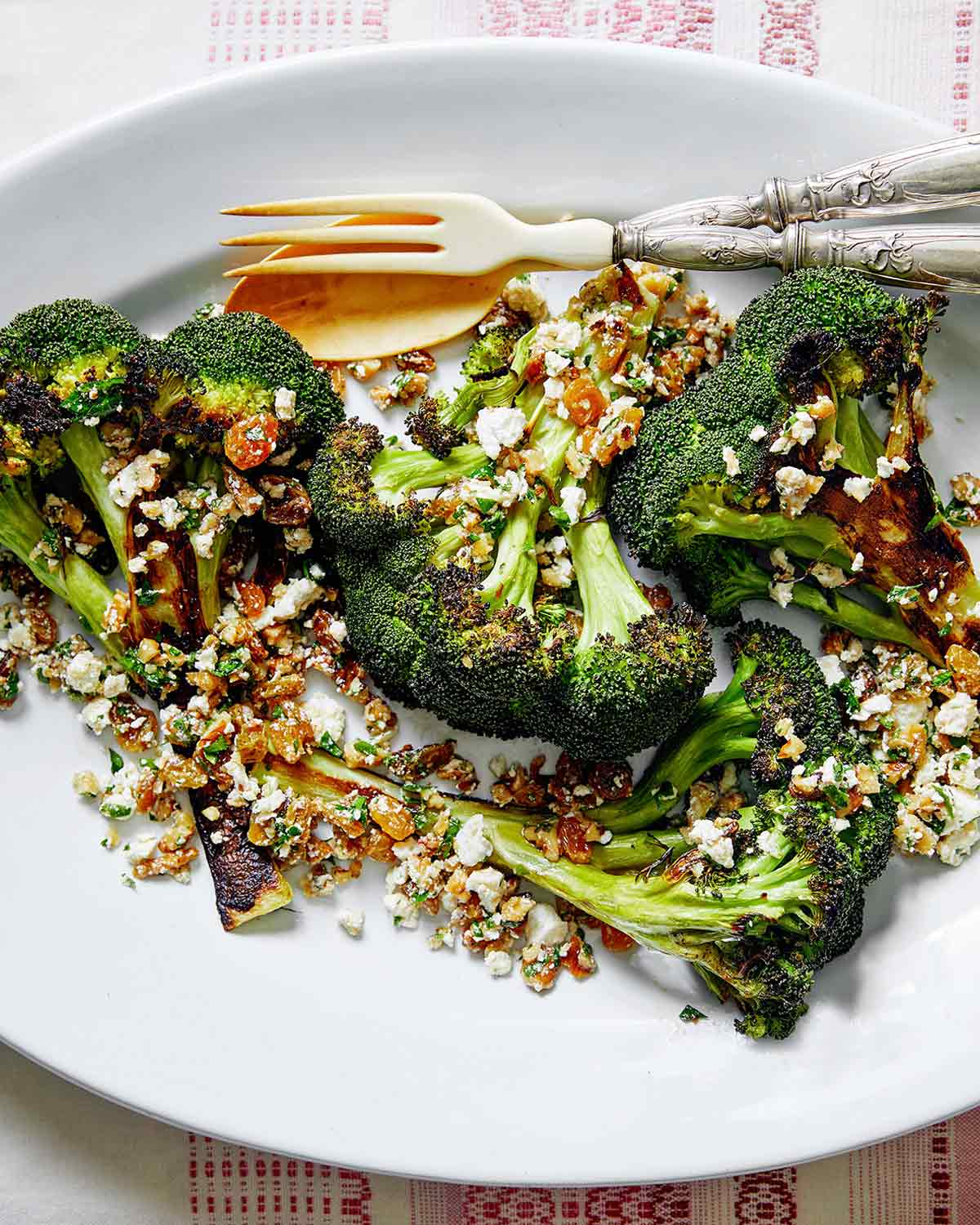 A white oval plate topped with broccoli with walnuts, raisins, and ricotta salata with a fork and spoon on the side.