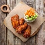 Five broiled buffalo wings on a piece of parchment on a wooden board with a bowl of carrot and celery sticks.