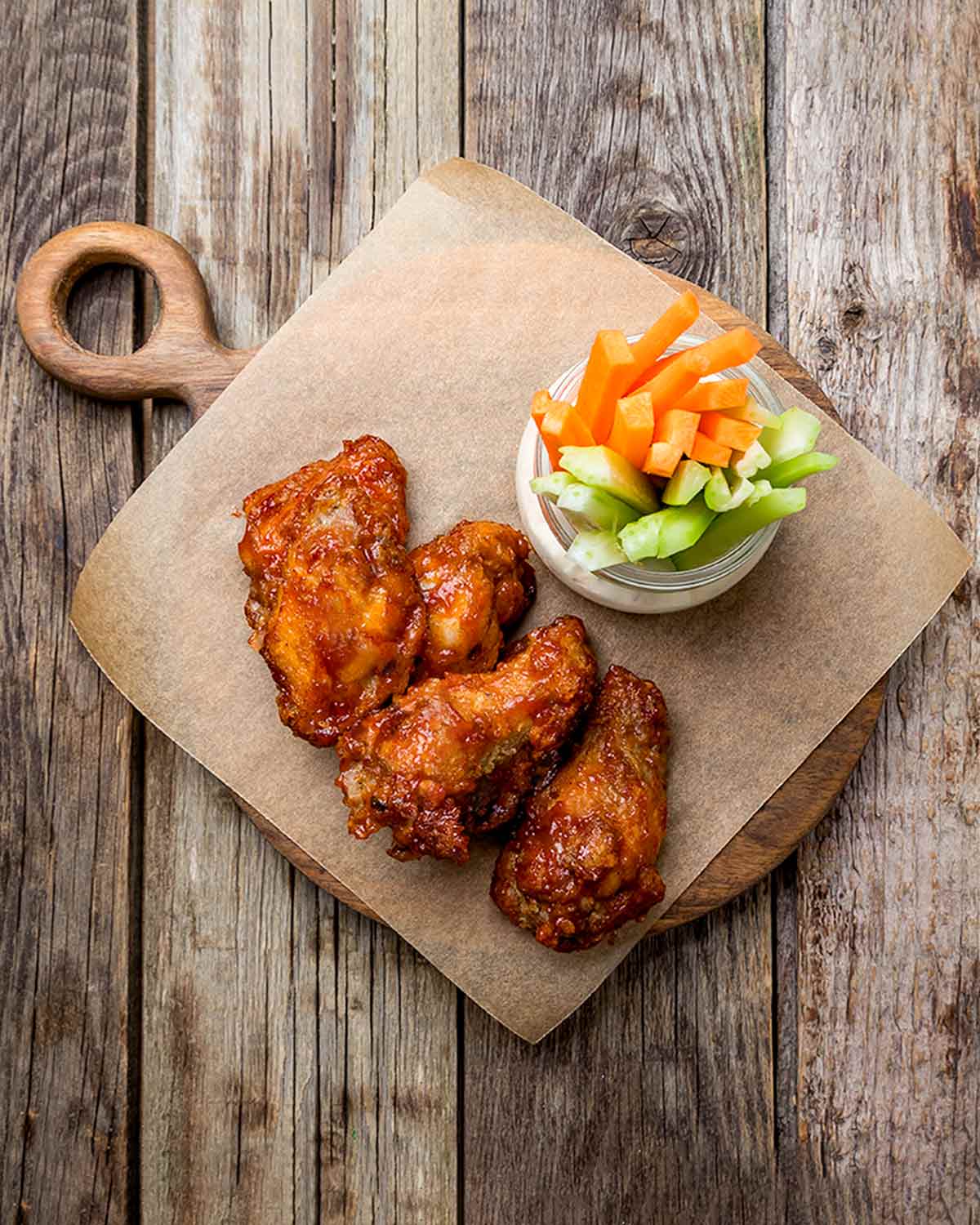 Five broiled buffalo wings on a piece of parchment on a wooden board with a bowl of carrot and celery sticks.