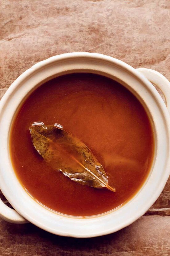 A white crock filled with brown vegetable stock with a bay leaf floating on top.