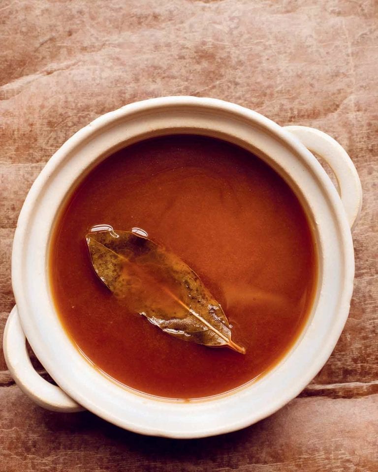 A white crock filled with brown vegetable stock with a bay leaf floating on top.