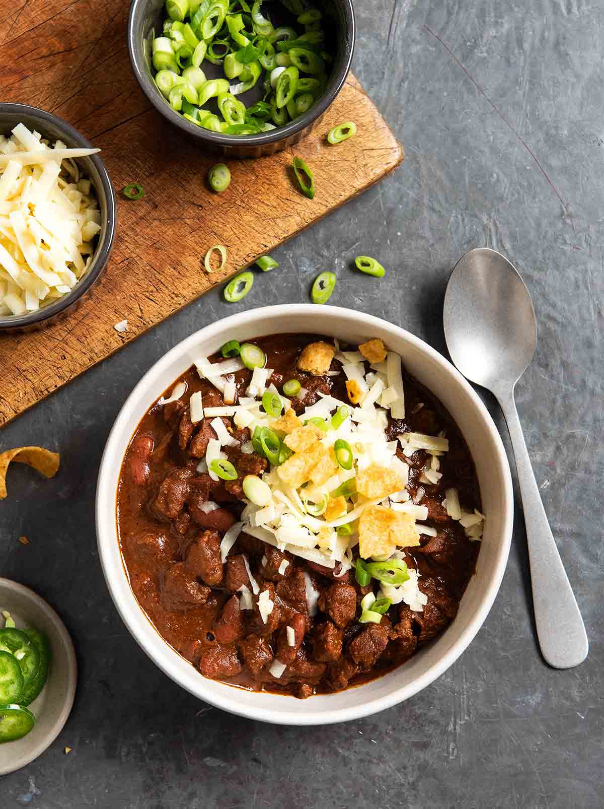 A bowl of chili con carne with beans, topped with cheese, scallions, and chips, with a spoon, a cutting board, and bowls of toppings on the side.