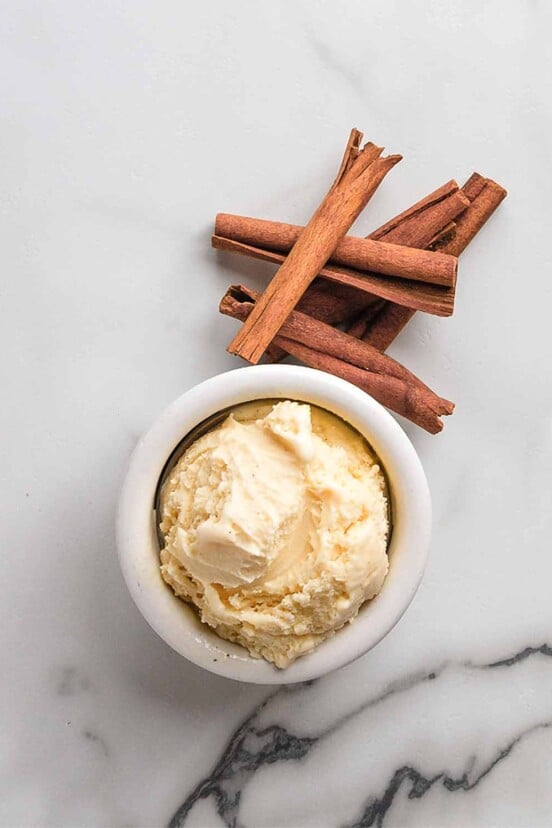 A small bowl of cinnamon gelato with cinnamon sticks next to it on white marble surface.