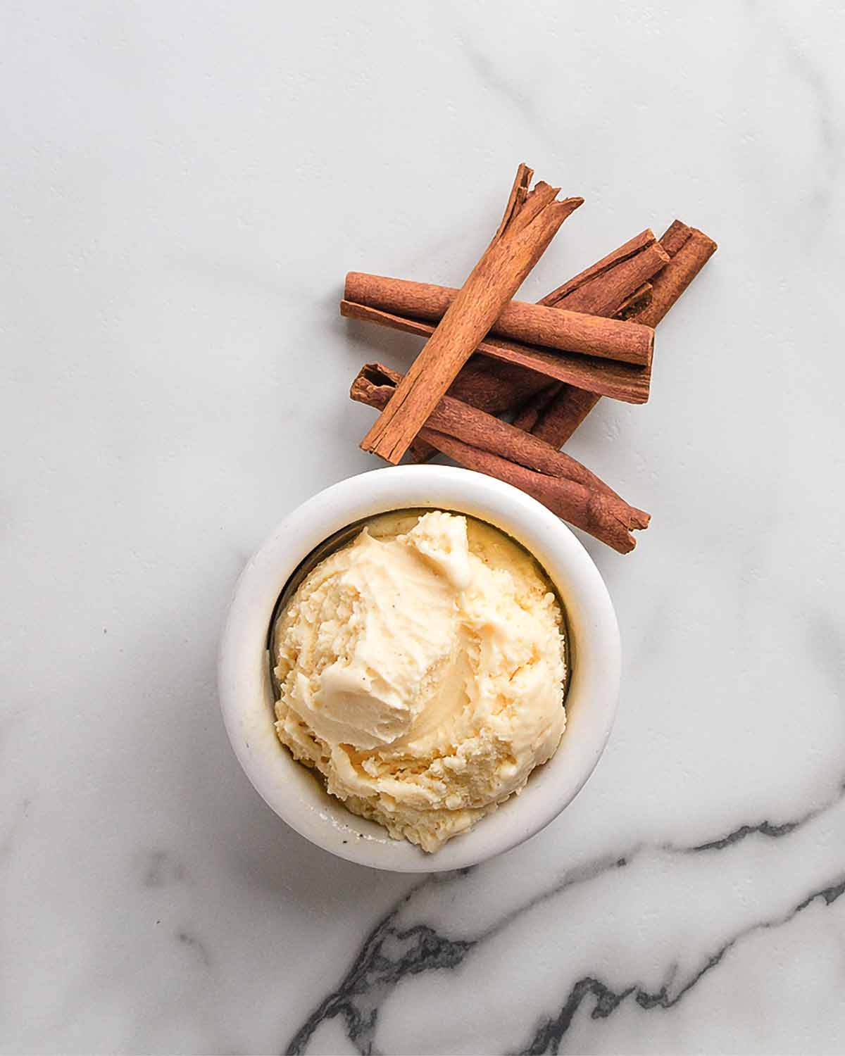 A small bowl of cinnamon gelato with cinnamon sticks next to it on white marble surface.