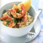 A white bowl of citrus risotto and garlic-chile prawns on a white plate with a fork on the side.