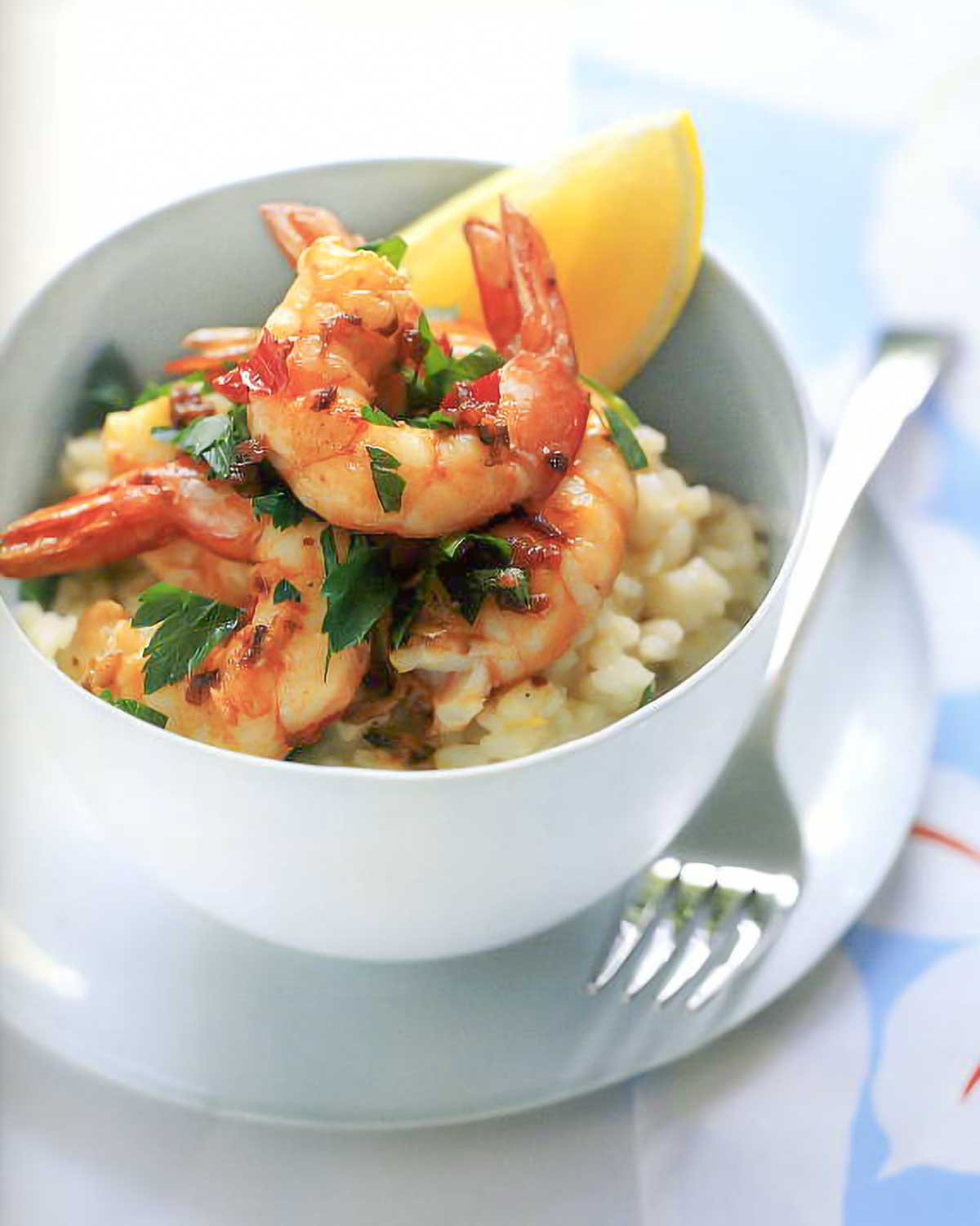 A white bowl of citrus risotto and garlic-chile prawns on a white plate with a fork on the side.