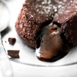 A molten chocolate cake on a plate with the filling oozing out and a few pieces of dark chocolate on the side.