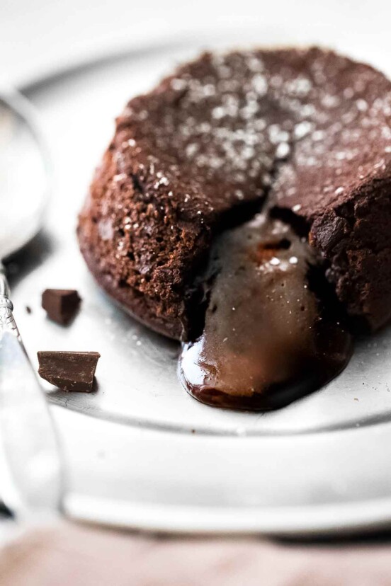 A molten chocolate cake on a plate with the filling oozing out and a few pieces of dark chocolate on the side.