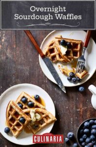 Two white plates with overnight sourdough waffles, blueberries, butter, and syrup on them.
