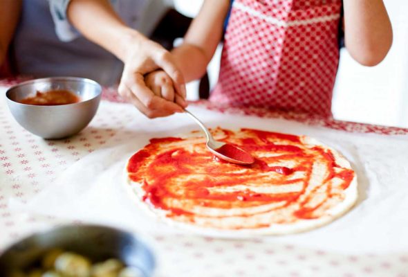 A person helping a child spread tomato sauce on a homemade pizza.