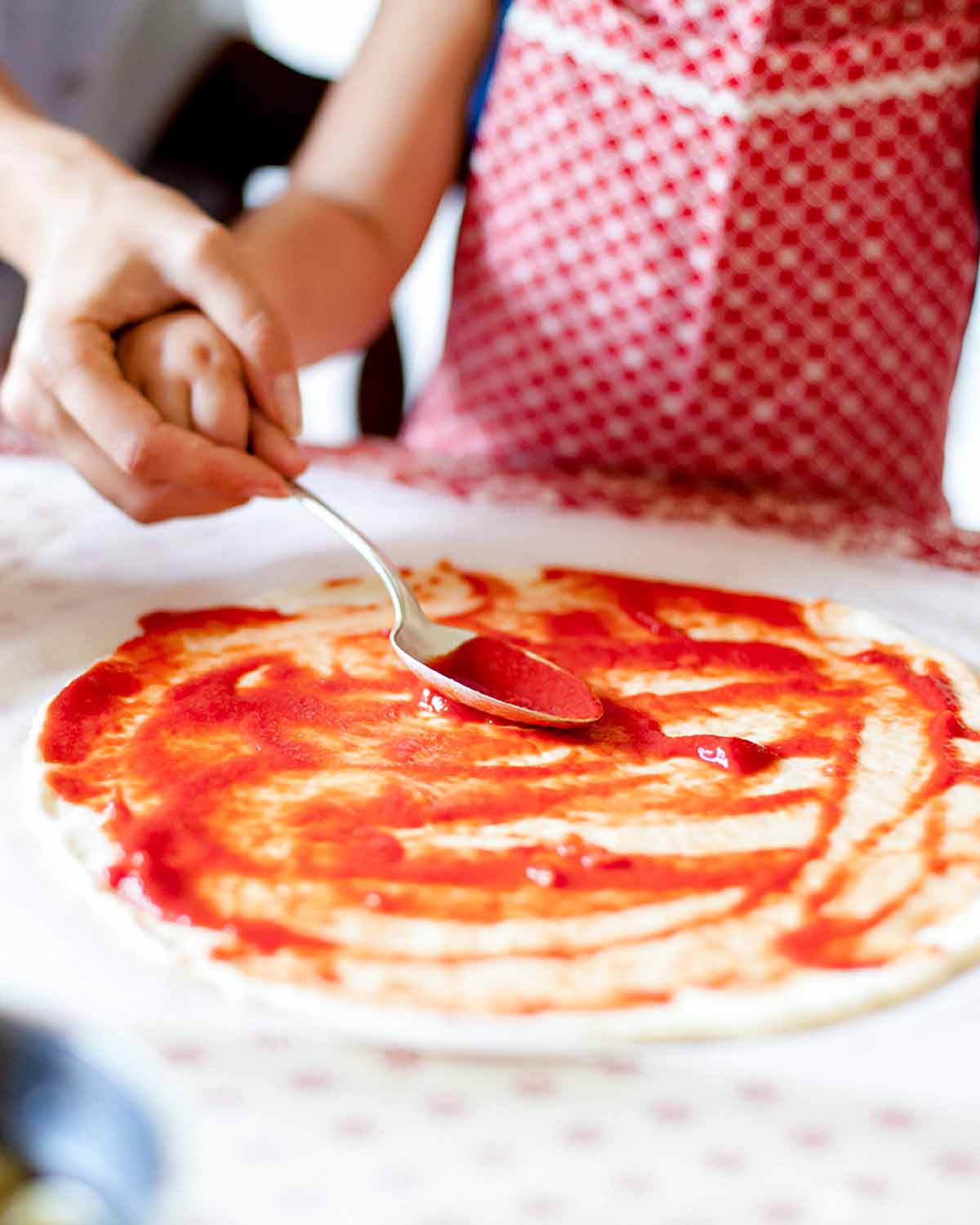A person helping a child spread tomato sauce on a homemade pizza.