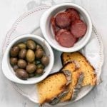 Slices of broa, or Portuguese corn bread, on a plate with a bowl of olives and an bowl of choiriço sliced
