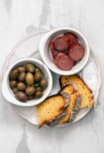 Slices of broa, or Portuguese corn bread, on a plate with a bowl of olives and an bowl of choiriço sliced.