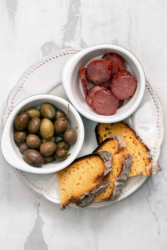 Slices of broa, or Portuguese corn bread, on a plate with a bowl of olives and an bowl of choiriço sliced.