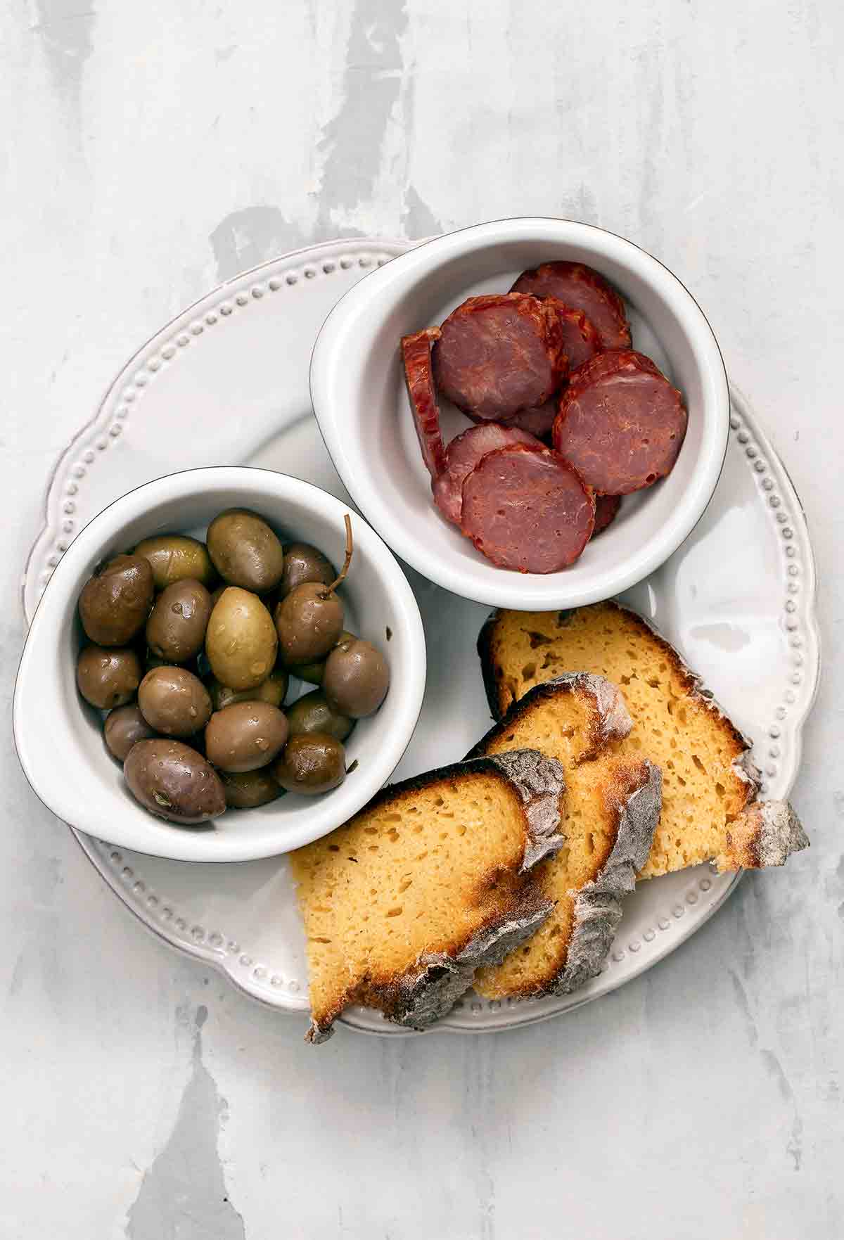 Slices of broa, or Portuguese corn bread, on a plate with a bowl of olives and an bowl of choiriço sliced