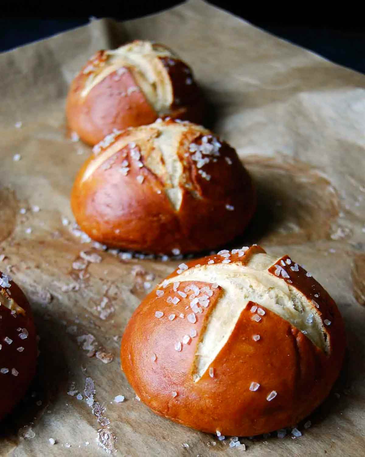 Three pretzel rolls on a sheet of parchment sprinkled with coarse salt.