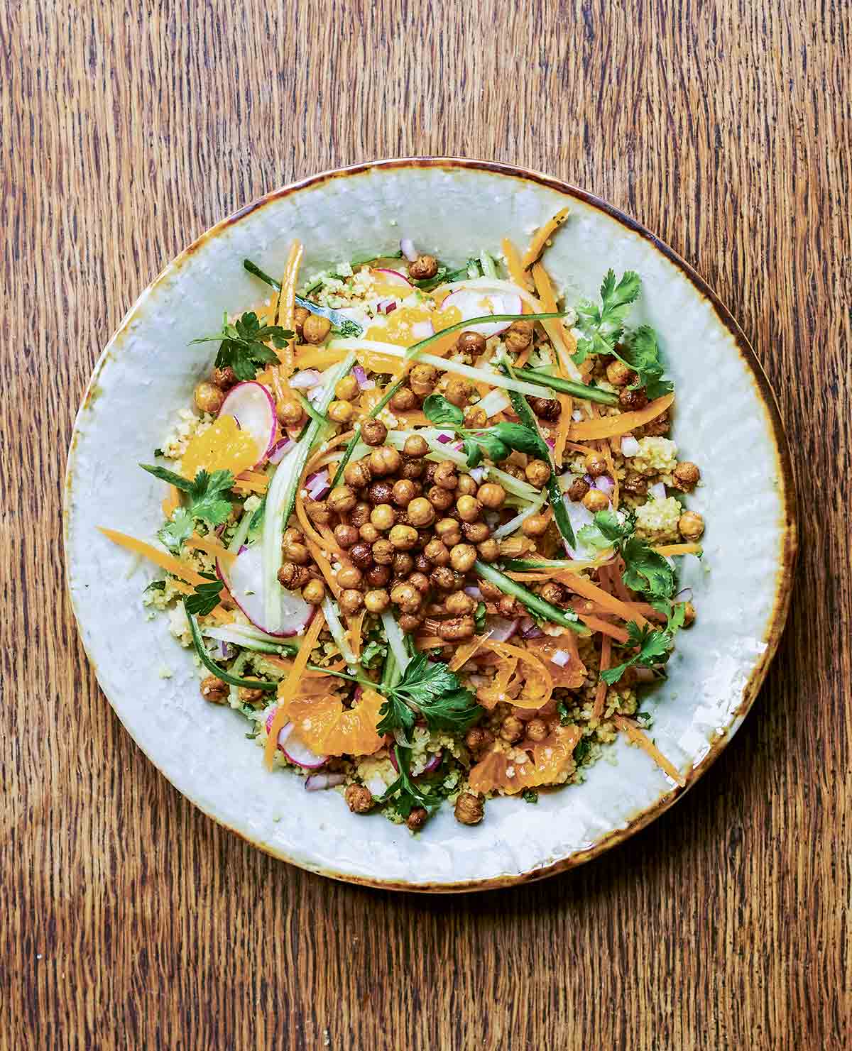 A gold-rimmed white plate topped with chickpea salad, orange segments, couscous, cilantro, and shaved carrot.