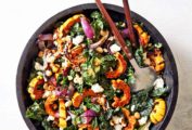 A black bowl filled with roasted delicata squash and kale salad with salad servers resting in it.