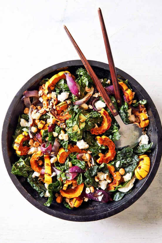 A black bowl filled with roasted delicata squash and kale salad with salad servers resting in it.