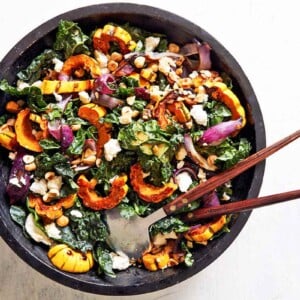 A black bowl filled with roasted delicata squash and kale salad with salad servers resting in it.