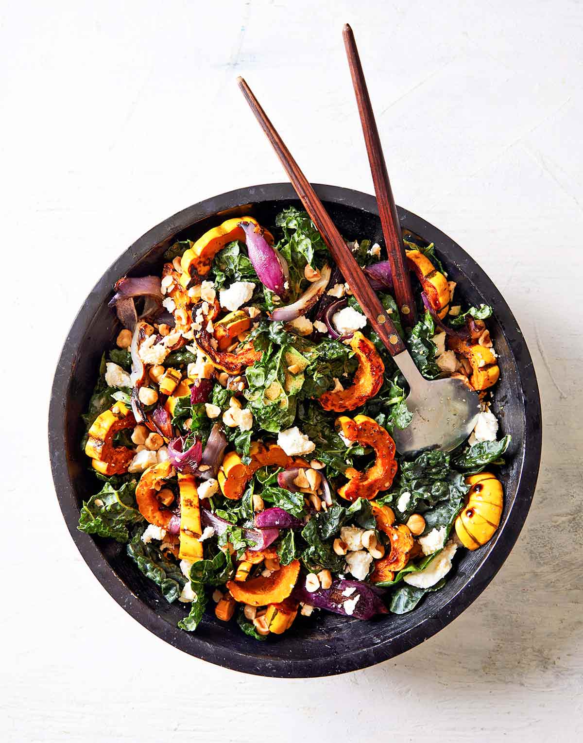 A black bowl filled with roasted delicata squash and kale salad with salad servers resting in it.