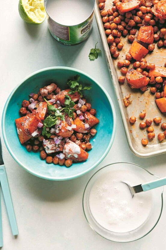 A bowl of roasted sweet potatoes and chickpeas, garnished with cilantro, next to a sheet pan and a bowl of coconut milk.