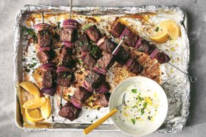 Sheet pan beef skewers piled on a foil-lined baking sheet with a dish of white sauce and lemon wedges.