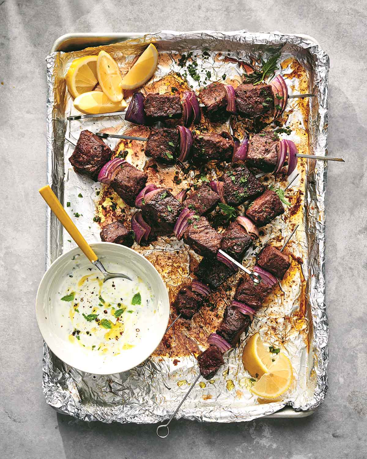 Sheet pan beef skewers piled on a foil-lined baking sheet with a dish of white sauce and lemon wedges.