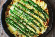 A cast-iron skillet filled with asparagus frittata on a wooden table with a fork on the side.