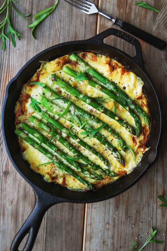 A cast-iron skillet filled with asparagus frittata on a wooden table with a fork on the side.