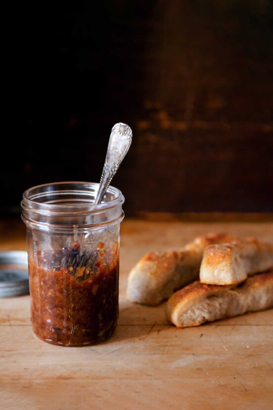 A jar of bacon jam with a spoon in it on a wooden board with a lid and breadsticks in the background.