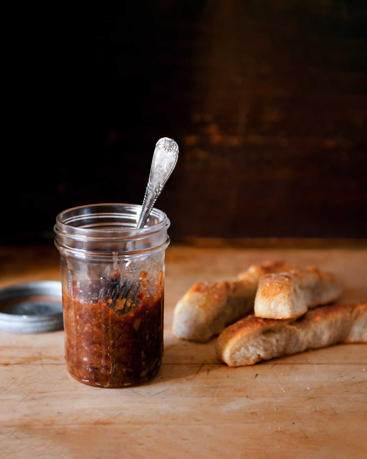 A jar of bacon jam with a spoon in it on a wooden board with a lid and breadsticks in the background.