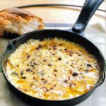 A cast iron skillet filled with bubbling baked fontina with garlic and thyme and a hunk of bread in the background.