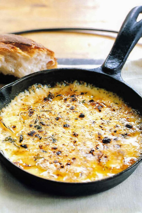 A cast iron skillet filled with bubbling baked fontina with garlic and thyme and a hunk of bread in the background.