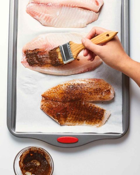 A person making baked tilapia with chile and lime by brushing tilapia fillets with a spice mixture.