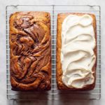 A loaf of banana bread with swirls of nutella next to a loaf of banana bread with cream cheese frosting on a wire rack.