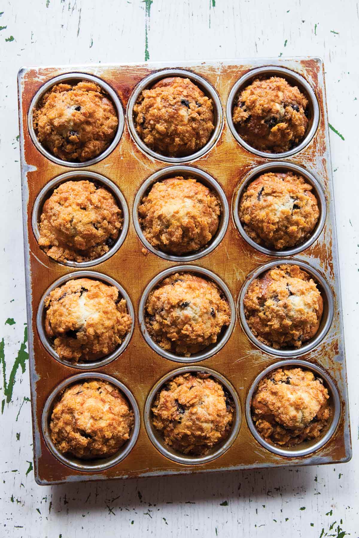 A 12 cup muffin tin filled with blueberry muffins with cornflake topping.
