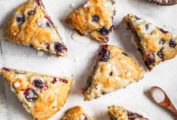 Eight blueberry scones on a piece of parchment with a dish of glaze and spoon lying nearby.