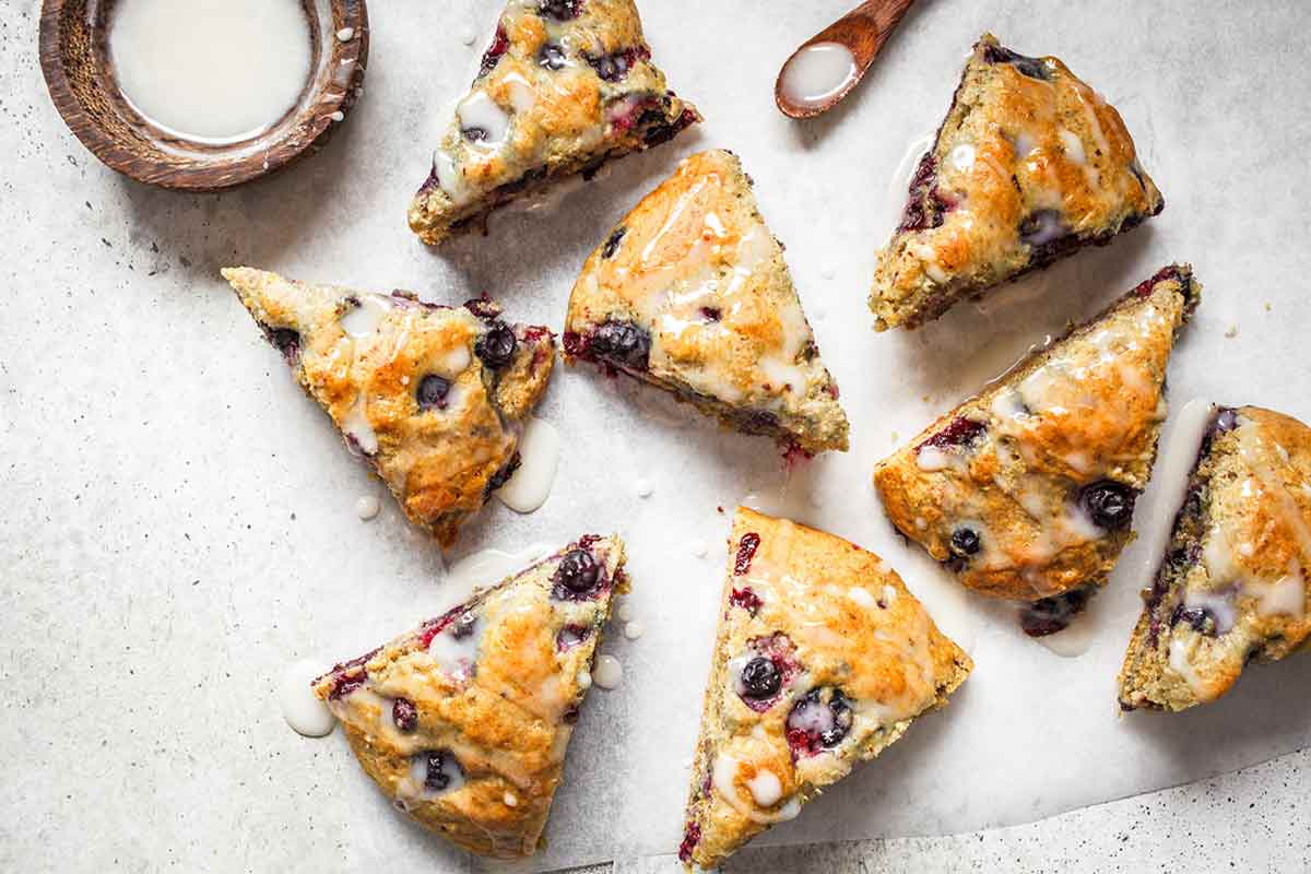 Eight blueberry scones on a piece of parchment with a dish of glaze and spoon lying nearby.
