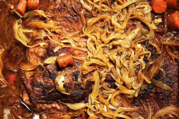 A partially submerged braised brisket with onions and carrots scattered on and around it.