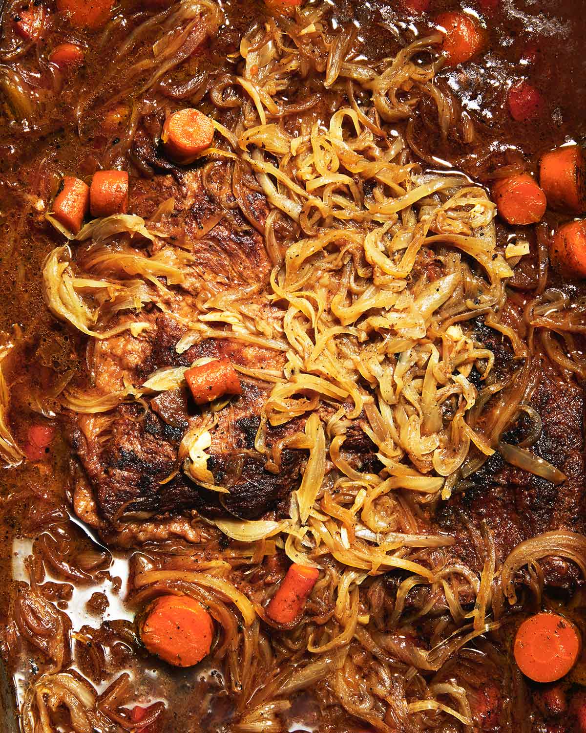 A partially submerged braised brisket with onions and carrots scattered on and around it.