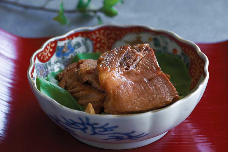 A patterned bowl with slices of braised pork with soy sauce on a red tray.