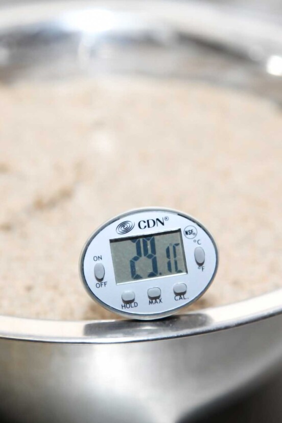 A digital thermometer sitting in bread dough that is proofing in a silver bowl.