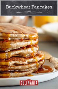 A stack of buckwheat pancakes on a white plate with maple syrup drizzled over them.