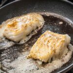Two pieces of butter basted fish with garlic and thyme in a skillet with butter foaming around them.