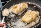 Two pieces of butter basted fish with garlic and thyme in a skillet with butter foaming around them.