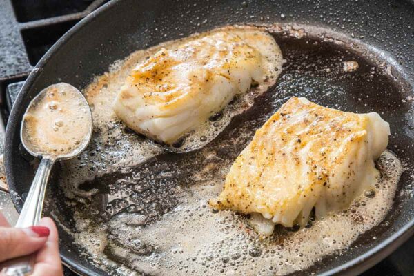 Two pieces of butter basted fish with garlic and thyme in a skillet with butter foaming around them.